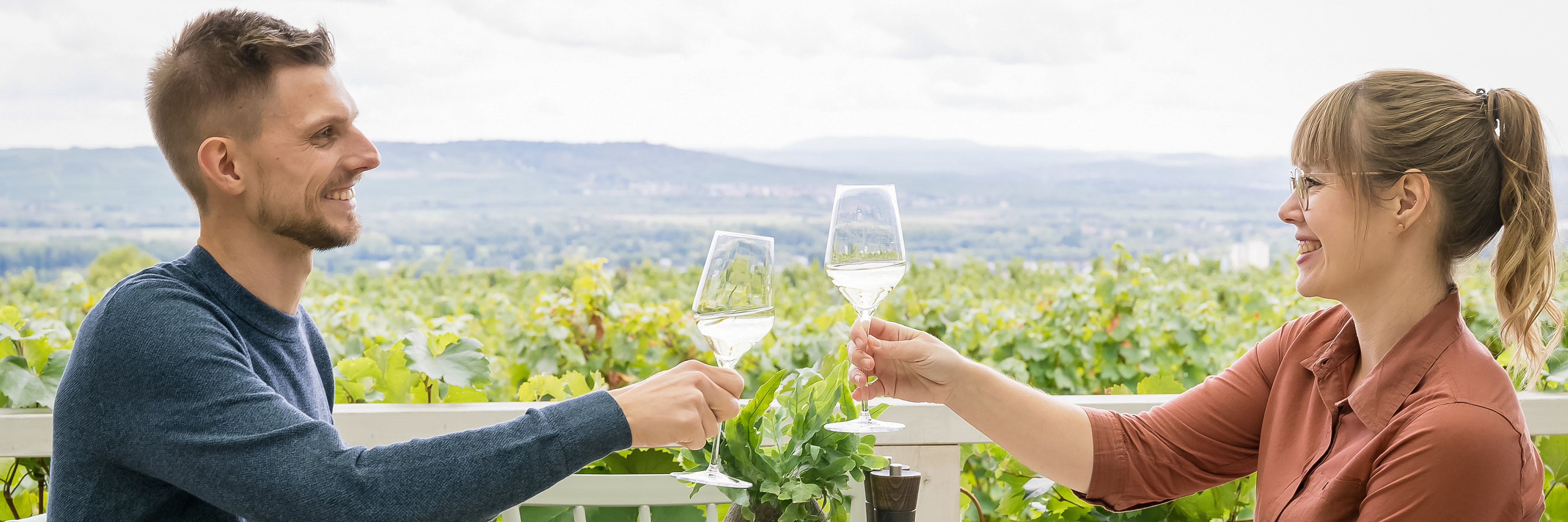 Kulinarik Ein Paar sitz auf einer Terrasse mit Blick auf die Weinberge und prostet sich mit Weißweingläsern zu. Auf den Tellern haben sie Rheingauer Spezialitäten, wie Spundekäse