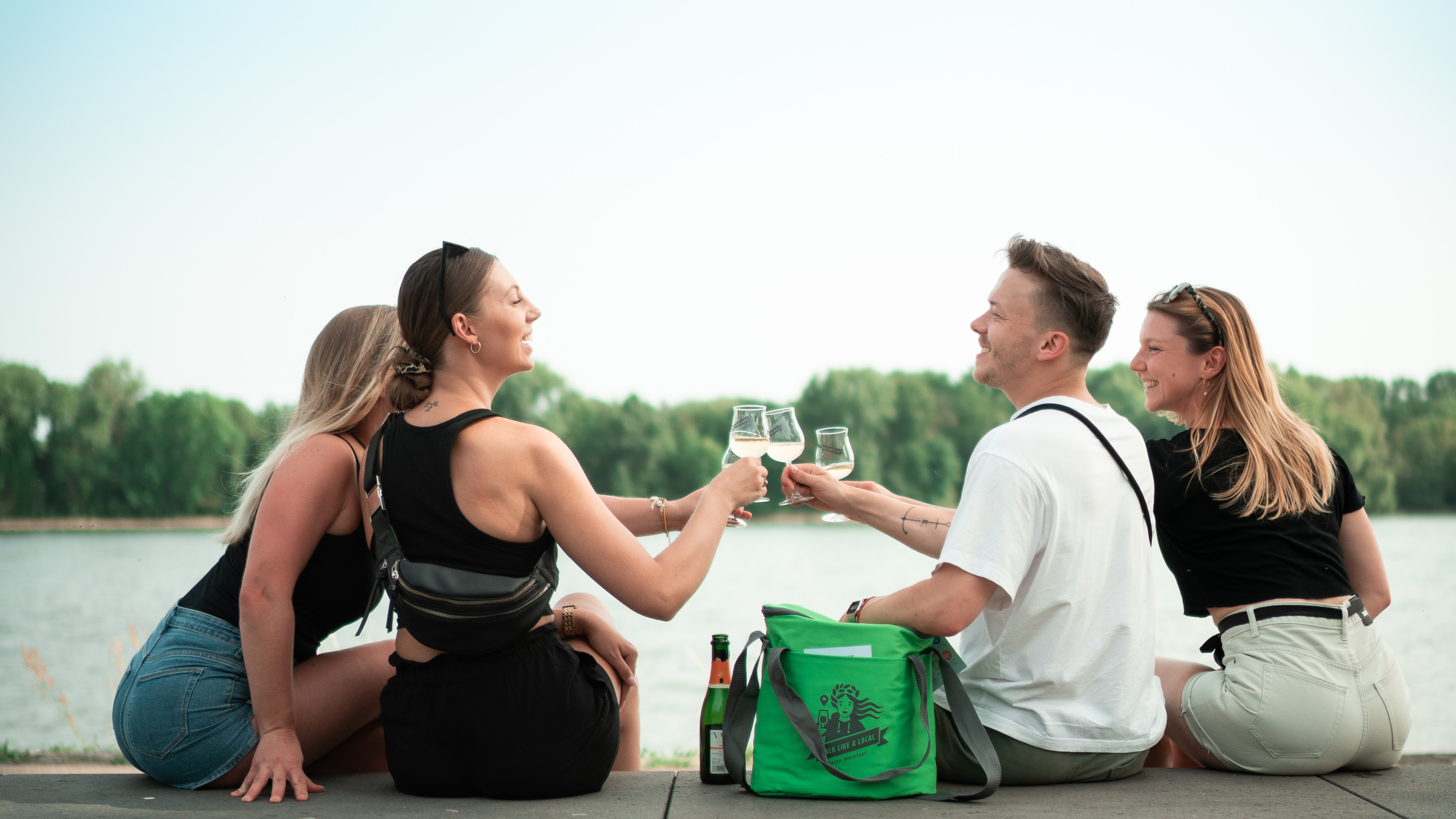 Vier Freunde sitzen am Rheinufer und stoßen mit ihren Walk like a local Weingläsern an. Zwischen ihnen steht die grüne Walk like a local Kühltasche.